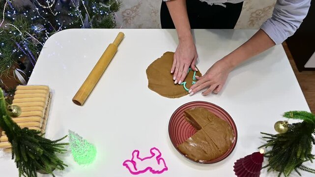 A girl cuts gingerbread dough using a man-shaped mold. The table is filled with baking tools and decorations. A Christmas tree and garland add to the festive mood of the setting.