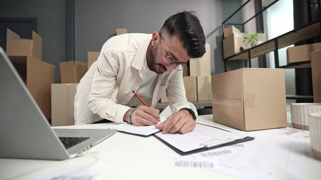 Man writes notes at desk with laptop and boxes nearby