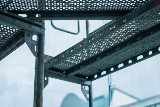 Perforated metal scaffold decks rest on tubular supports and connecting brackets, showing the underside of temporary platforms used for facade access and work at height