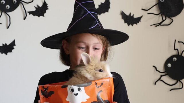 Video portrait happy excited face girl child in black witch hat holding orange candy bucket rabbit against a wall decorated background with bats, celebrating Halloween with playful costume. spooky han