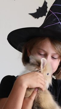 Video vertical  portrait happy excited face girl child in black witch hat holding orange candy bucket rabbit against a wall decorated background with bats, celebrating Halloween Pet shop advertising