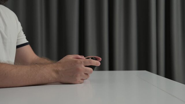 Close up hands holding wired black game controller over glossy white tabletop, thumbs poised above buttons, cable trailing toward gray curtain backdrop in indoor studio