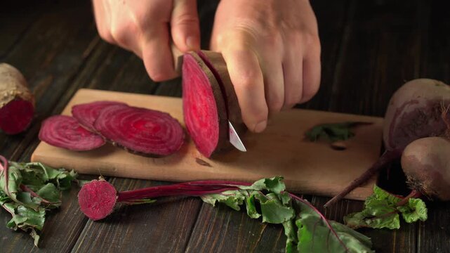 Hands of a person slicing a fresh beetroot on a wooden cutting board, with vibrant red beet slices and leafy greens arranged around, creating a rustic kitchen atmosphere