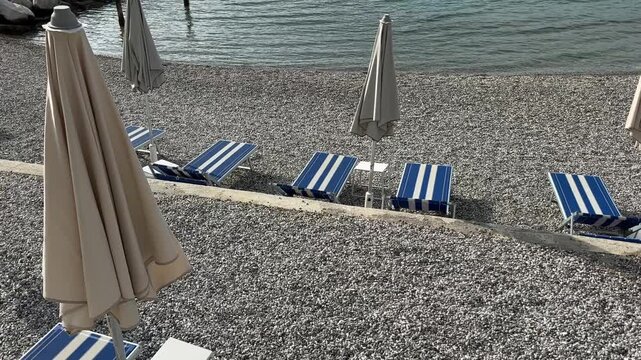 High angle view of empty blue striped loungers with folded sun umbrellas placed on pebble beach near calm sea water during sunny morning.