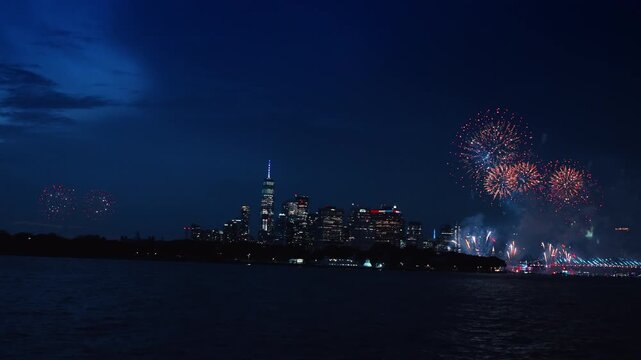 New York downtown skyline with double firework display over river.. Bright pyrotechnics explode on both sides of illuminated skyscrapers reflecting on dark water surface at night.