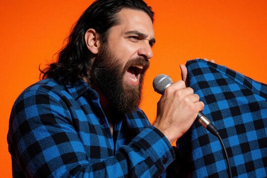 Bearded man sings loudly into microphone. He wears a blue checkered shirt against an orange background, showing passion and energy. Male performer expresses emotions while singing song.