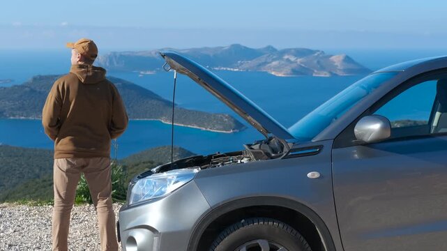 Broken car on a trip. Frustrated man standing on a scenic road looking at his broken-down car with an open hood during a road trip with a beautiful sea view
