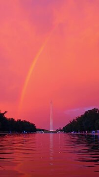 Red sunset sky and rainbow over Washington Monument.. The stone obelisk reflects in a long water pool surrounded by dark trees under a bright crimson evening atmosphere.