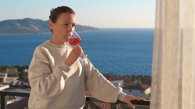 Relaxed woman drinking wine on balcony with sea view. Content woman enjoying a glass of red wine on a balcony, savoring the moment while looking out at the beautiful blue sea and coastal town