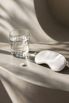 Minimalist healthcare still life with white tablet, glass of water and sleep mask on a sculptural surface, symbolizing medication, sleep, insomnia treatment and nighttime wellness.