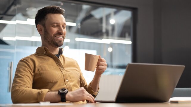Smiling Caucasian Man In His 30s Enjoying A Cup Of Coffee While Working On Laptop At Modern Office Workspace. Engaged In Professional Tasks, Representing Business Concept.