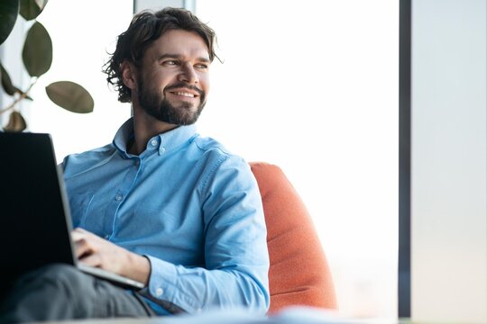 Smiling Caucasian Man In His 30s Working On Laptop While Sitting In A Bright Modern Office. Focused On Task, Enjoying Productive Day In Professional Environment. Business Concept.