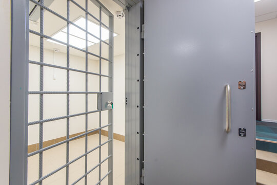 A doorway of a prison cell is visible with a metal door opened partially. The inside of the secure facility appears clean and well-lit. The floor and walls are neutral in color