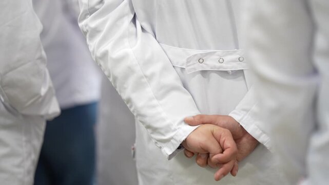 Worker holds one wrist behind a work coat with a snap fastened back belt, showing garment construction and standing posture in a group wearing similar uniforms