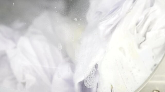 Laundry moves inside a drum behind glass carrying soap bubbles and streaks, illustrating mechanical agitation of fabric during commercial machine washing