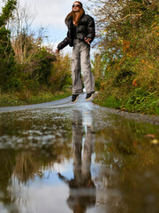 Woman stands on wet road, reflection in a puddle visible. Autumn leaves surround path. Trees and sky reflect in puddle. Neutral tones and natural lighting. Selective focus. Young teenager outdoor. © mark_gusev