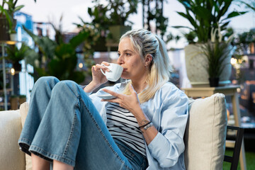 Obraz premium Woman drinks coffee from a white cup while relaxing on a rooftop garden sofa, enjoying a quiet leisure moment among plants in an urban setting.