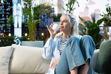 Obraz premium Woman drinks coffee from a white cup while relaxing on a rooftop garden sofa, enjoying a quiet leisure moment among plants in an urban setting.