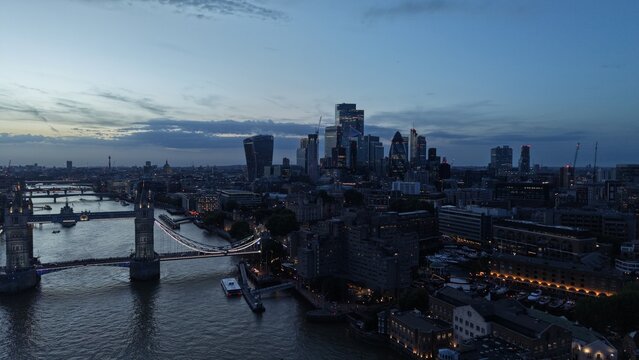 Aerial view of the City of London financial district at blue hour, with modern skyscrapers rising behind Tower Bridge and the River Thames as evening lights illuminate the city.