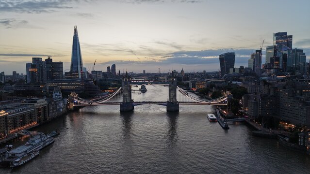 Symmetrical aerial view of Tower Bridge over the River Thames at dusk, framed by The Shard and the modern City of London skyline with illuminated buildings and boats on the river.