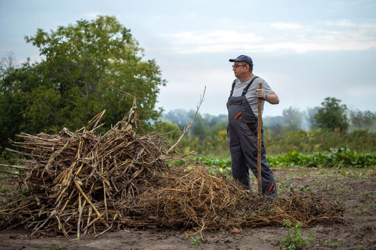 Field worker stands beside haylike pile holding shovel. Adult man in work overalls pauses at muddy rural plot, near dried crop remains, with calm determined expression and practical outdoor setting.
