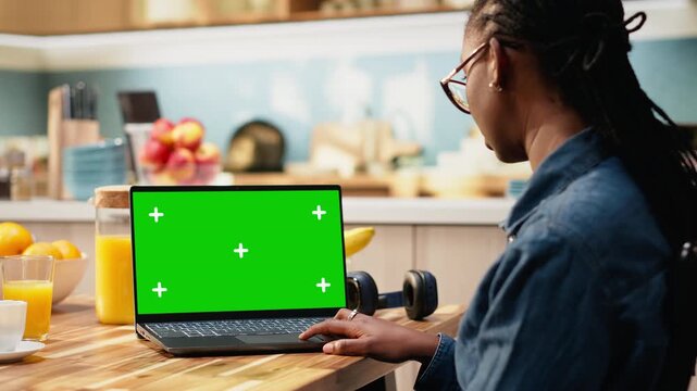 Woman works from home in kitchen with isolated chroma key on laptop, focused on editable replaceable mockup screen. Freelancer lifestyle highlights productivity during daytime remote work at home.