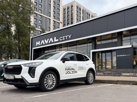 White Haval Jolion test drive SUV car parked outdoors on asphalt street next to a modern Haval City dealership showroom glass building facade. Premium Chinese crossover automobile transport vehicle. 