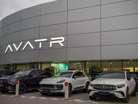 Exterior facade of a modern Chinese Avatr car dealership building with corporate lettering logo. Premium Mercedes-Benz and Porsche SUV automobiles parked outdoors near a glass showroom pavilion.