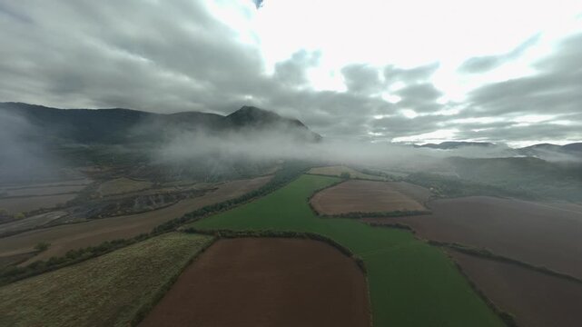 High FPV flight above geometric green and brown farm fields in a broad valley with sunlight breaking through layered storm clouds