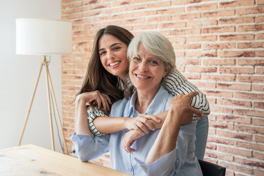 Cheerful senior mother and adult daughter embrace and smile at the camera in a warm indoor setting, sharing a loving family moment of connection, care, and joy