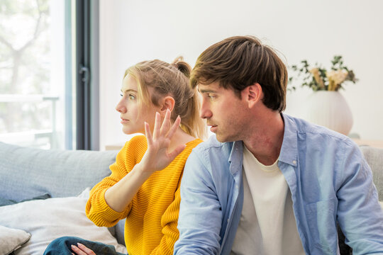 A young couple sits on a sofa during a tense moment, the woman turning away and raising her hand while the man looks upset and distant, conveying conflict, rejection and relationship strain.