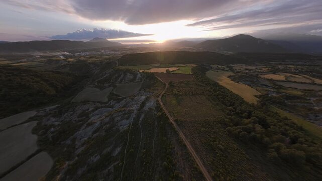 Wide FPV flight above rolling fields and winding tracks in a rural mountain valley with the setting sun glowing beneath layered clouds