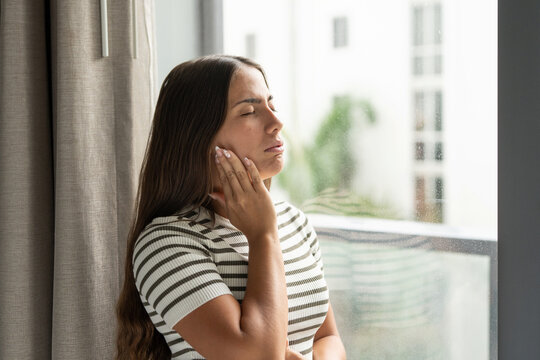 Young woman indoors clutching her cheek with eyes closed, wincing from severe toothache and jaw pain, showing oral discomfort and urgent need for dental care and relief