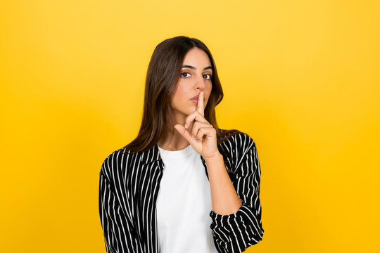 Woman stands against vibrant yellow background, finger to lips in a shushing gesture conveying secrecy, quiet, confidentiality and a request for silence or discretion