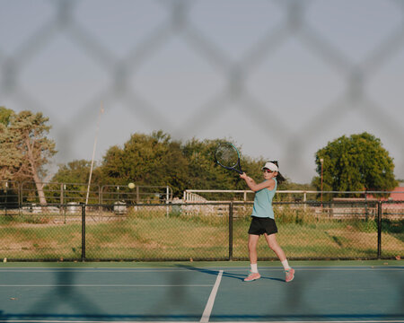 Young girl swinging a backhand stroke on an outdoor tennis court seen through a fence