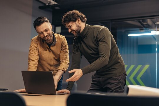 Smiling Male Professionals Engaging In Collaborative Work On Laptop In Contemporary Office Setting. Two Young Men Discussing Ideas And Strategies For Project Implementation Together.