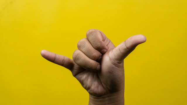 One adult male hand demonstrates relaxed shaka signs extending thumb and pinky outward against a vivid yellow background. Remaining three central digits curl tightly toward palm