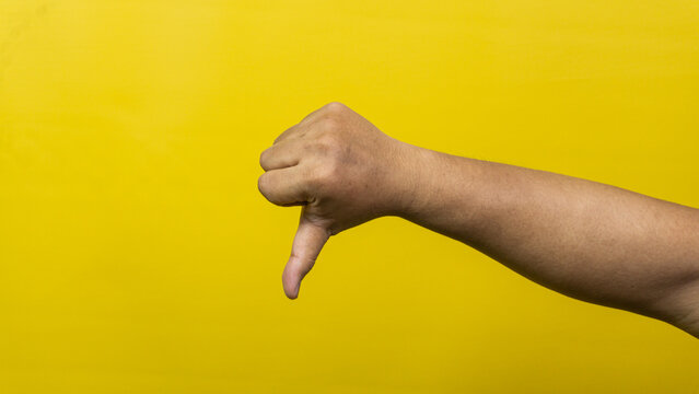 A right arm is extended horizontally, displaying a thumbs down gesture against a solid, vibrant yellow background. The hand is closed in a fist with the thumb pointing straight down