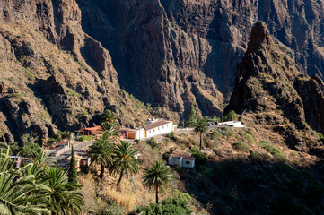 A long white farmhouse stands on a rocky spur below the sheer walls of the Masca gorge. Remote rural property and mountain living for travel and lifestyle storytelling. © Oksana