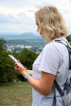 Vertical photo of a blonde woman looking at her smartphone while hiking. Female tourist with a backpack using a navigation app outdoors. City and mountains in the background
