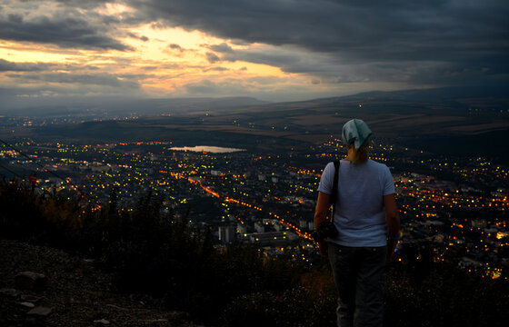 Rear view of a female photographer looking at a city at dusk. Woman standing on Mashuk mountain top overlooking illuminated urban landscape during sunset. Travel and night concept