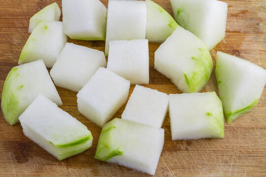 Raw cut winter melon pieces, with green skin patches, placed on a wooden cutting board, prepared for cooking.