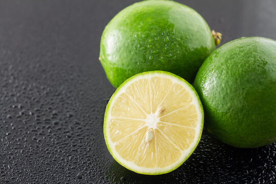 Whole and sliced fresh limes with water droplets, placed on a dark surface, showing juicy flesh and green peel.