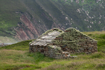 An old traditional stone croft house with a moss-covered roof sitting in a grassy valley with a steep rocky mountain in the background, Isle of Hoy. © Raisa