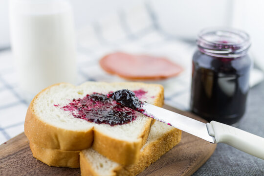 A knife spreads dark berry jam on a slice of white bread, with a jam jar and milk bottle nearby on a wooden board.