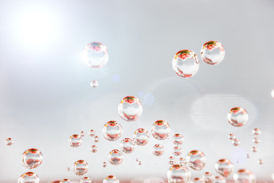 Close-up view of transparent, glossy water droplets of varying sizes suspended against a light neutral background, showcasing their smooth spherical shapes and subtle reflections.