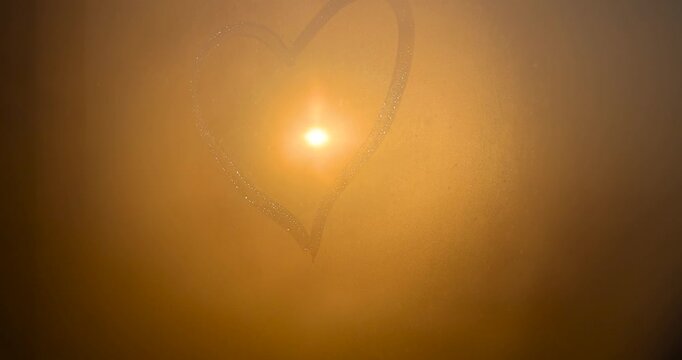 The golden disk of the sun outside the window with drops of water and a heart pattern on the glass, the bright sun illuminates small raindrops on the transparent surface