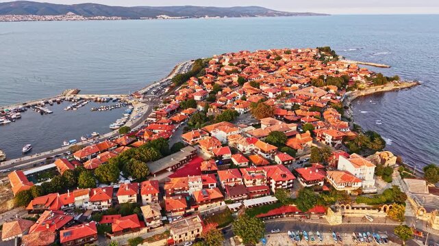 Houses with red roofs are seen along a narrow strip of land by the water. Boats are docked in the harbor. The scene shows a calm time of day.