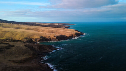 Aerial view of a coastline with blue ocean, waves hitting rocky cliffs, and dry hills under sky © HABI ANGGARA PUTRA