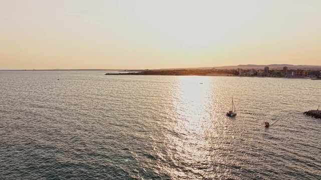 A boat floats on the water as the sun sets behind the shore. The coastline is visible in the distance, with clear skies and gentle waves.
